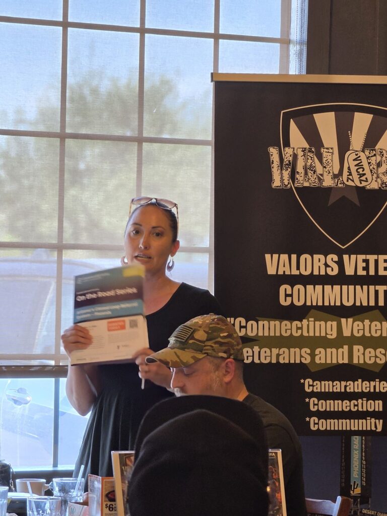 A woman holds up a flyer titled On the Road Series while speaking, with the VVCAZ banner behind her and attendees listening.