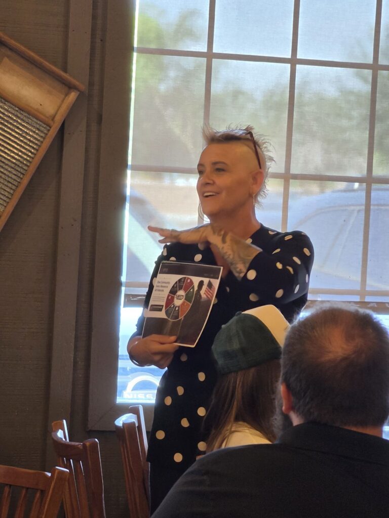 A smiling woman in a polka-dot top speaks while holding a brochure that reads One Community. Every Resource. All Veterans.