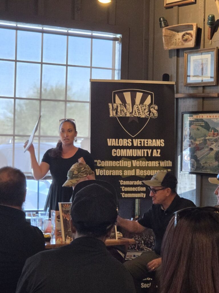 A woman in a black dress holds up a paper and speaks to the room in front of the Valors Veterans Community AZ banner while attendees listen.