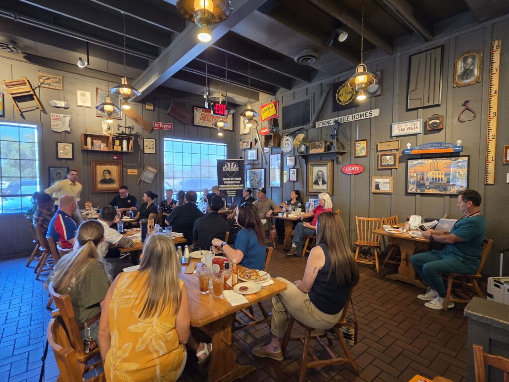 Wide view of a VVCAZ Coffee Connections gathering: veterans, family and community partners seated at tables in a restaurant dining room, with the Valors Veterans Community AZ banner at the back.
