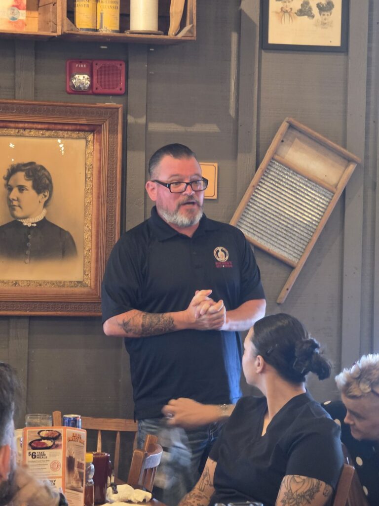 A man in a Soldier's Best Friend polo stands with his hands clasped and speaks to the group.