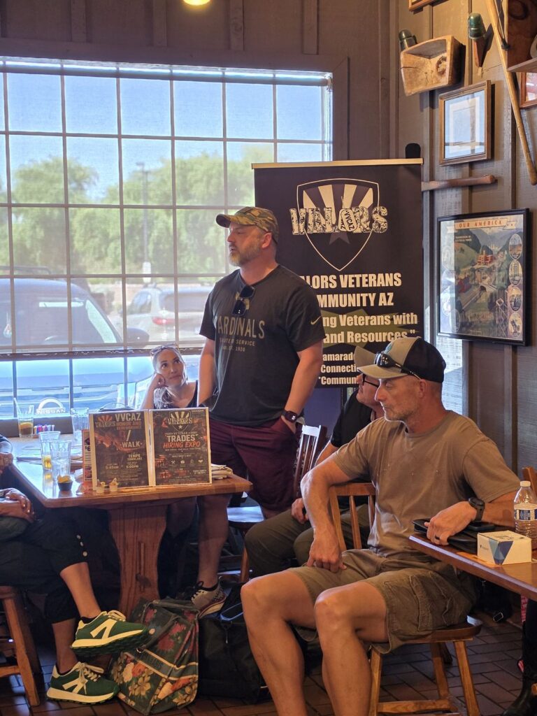 A man in a camouflage cap and a Cardinals Salute to Service shirt stands to speak to the group, with the VVCAZ banner and flyers for the Honor and Remembrance Walk and the Veteran Trades Hiring Expo behind him.