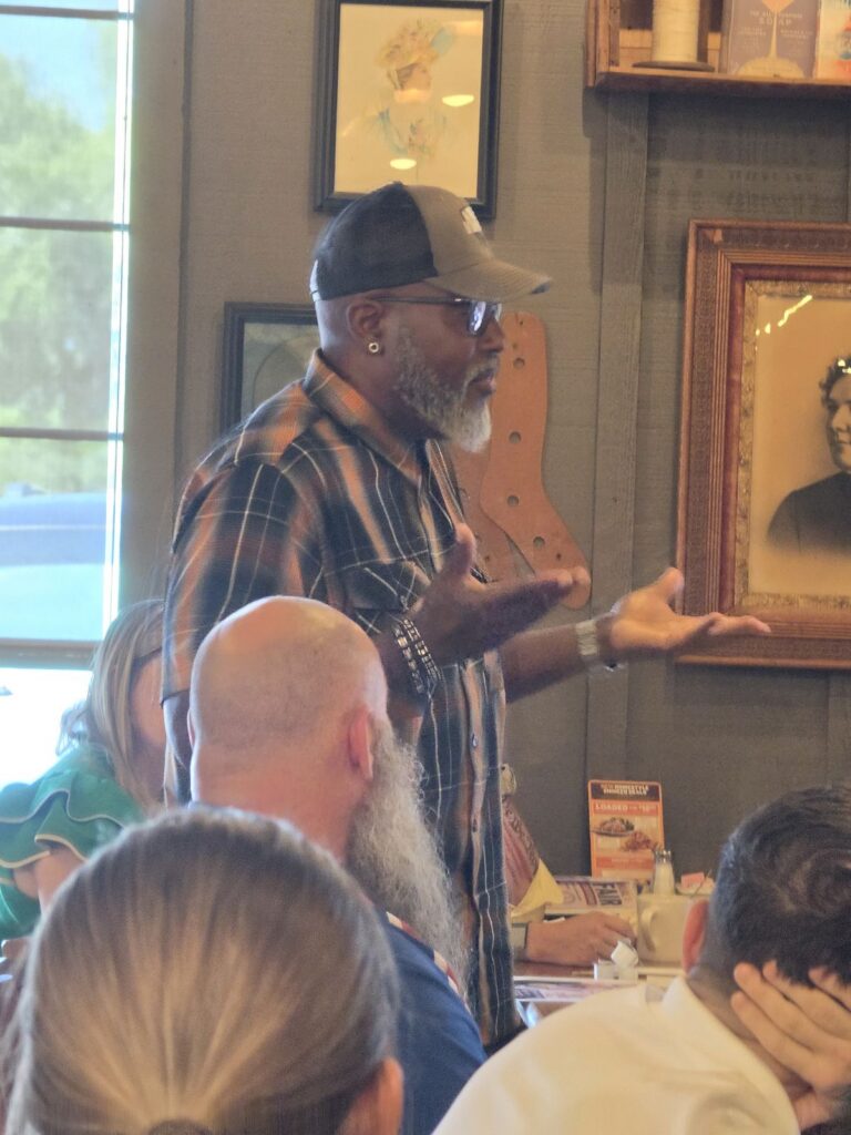 A man in a plaid shirt and cap gestures with open hands as he speaks to the group.
