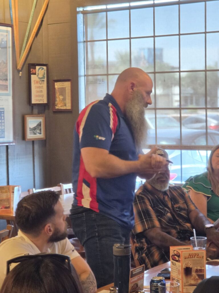 A bearded man in a red, white and blue polo stands to speak while seated attendees listen.