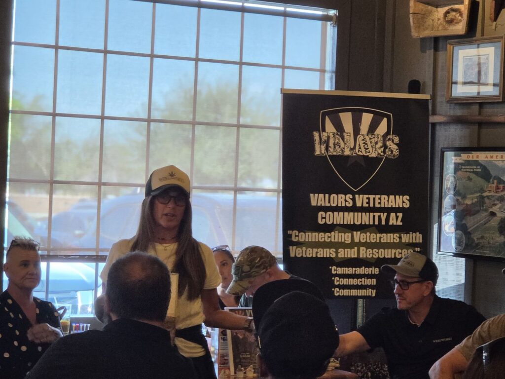 A woman in a trucker cap and glasses speaks to the room in front of the VVCAZ banner while seated attendees listen.