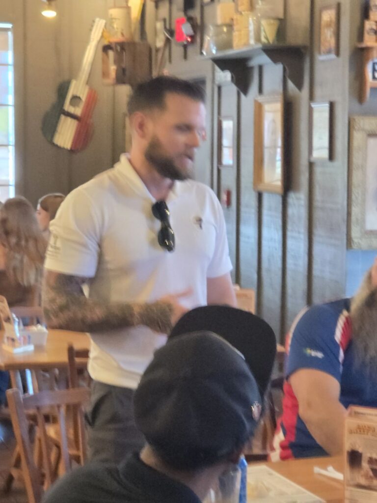 A man in a white polo shirt speaks to the group while seated attendees listen.
