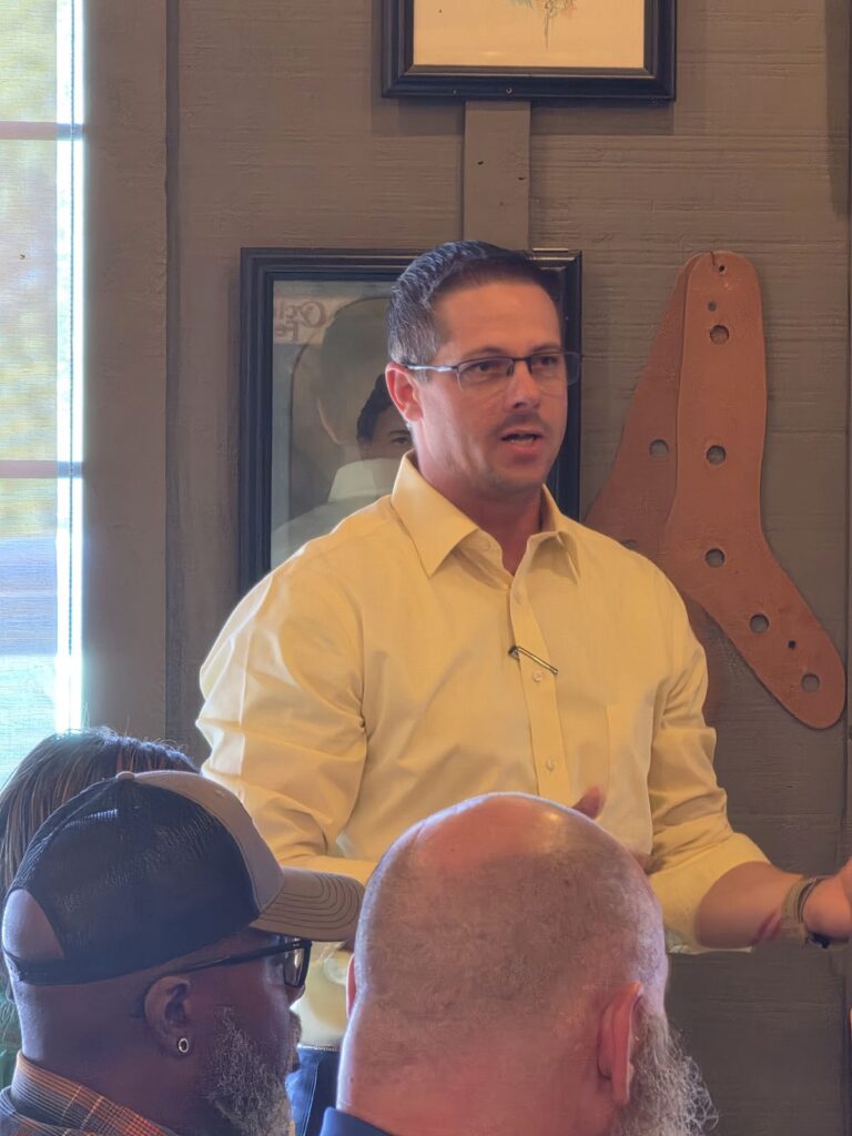 A man in a yellow button-up shirt speaks to the room while seated attendees listen.