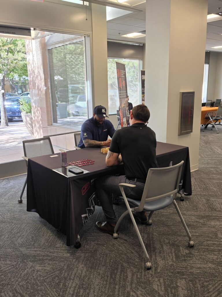Two men sit across from each other in conversation at an employer table, with other employers' banners behind them.