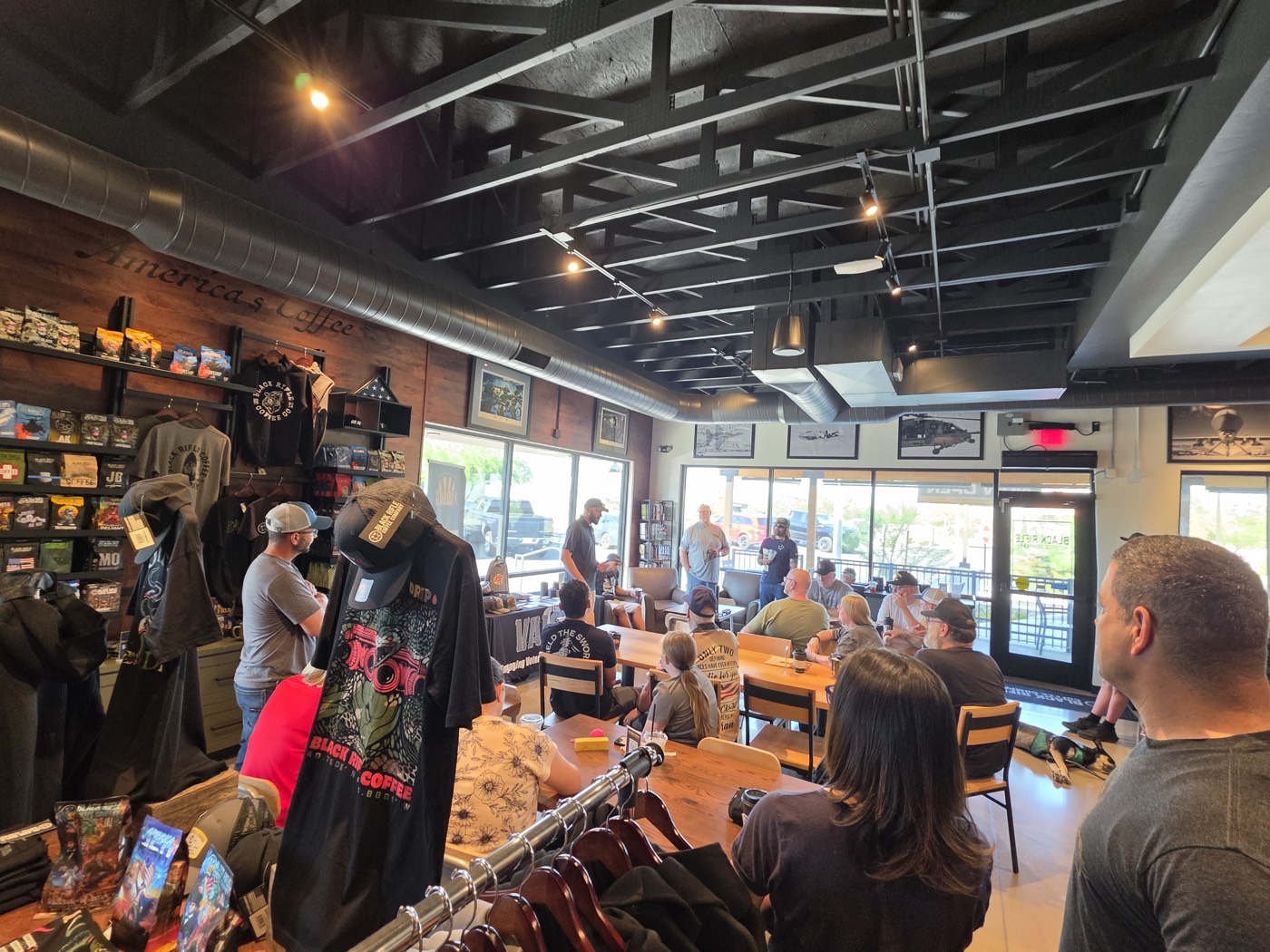 Attendees seated around wooden tables inside Black Rifle Coffee in Tucson, with shirts on a rack in the foreground, listening during Coffee Connections.
