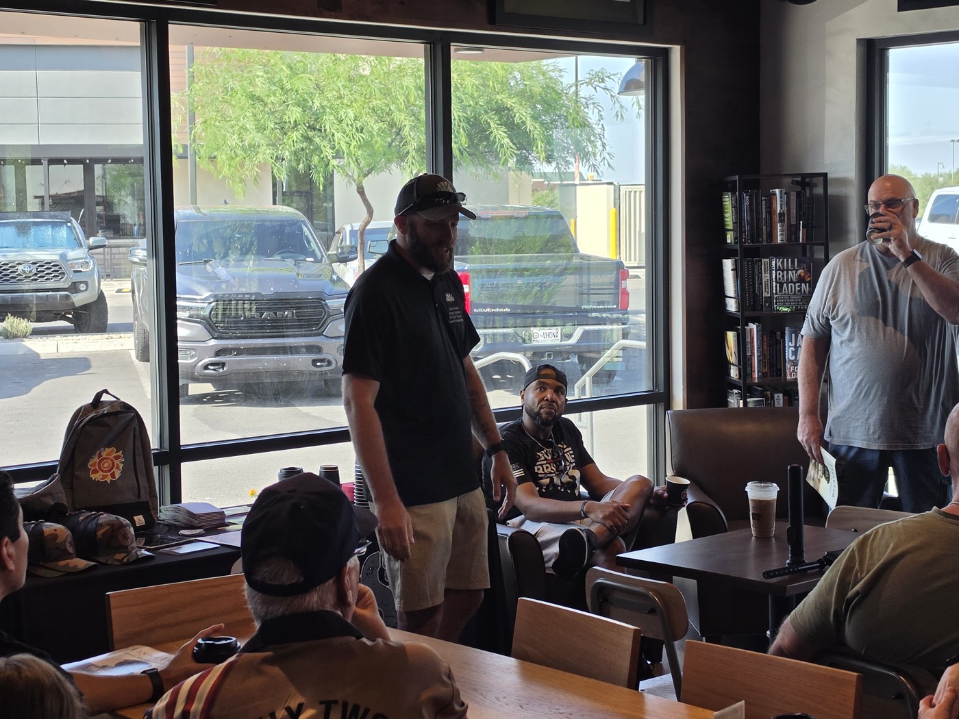 A bearded man in a black polo stands to speak to the group, with other attendees listening and the VVCAZ table visible at the left.