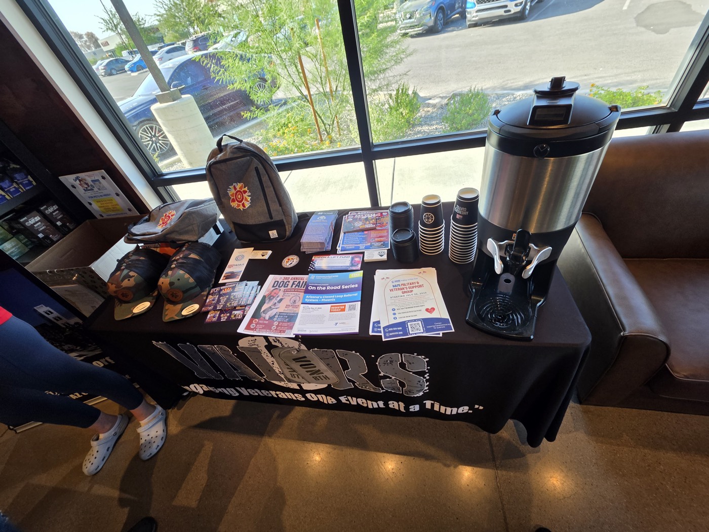 The VVCAZ table at Black Rifle Coffee in Tucson with a black Valors tablecloth, hats, a backpack, printed flyers including On the Road Series, and a coffee dispenser with paper cups.